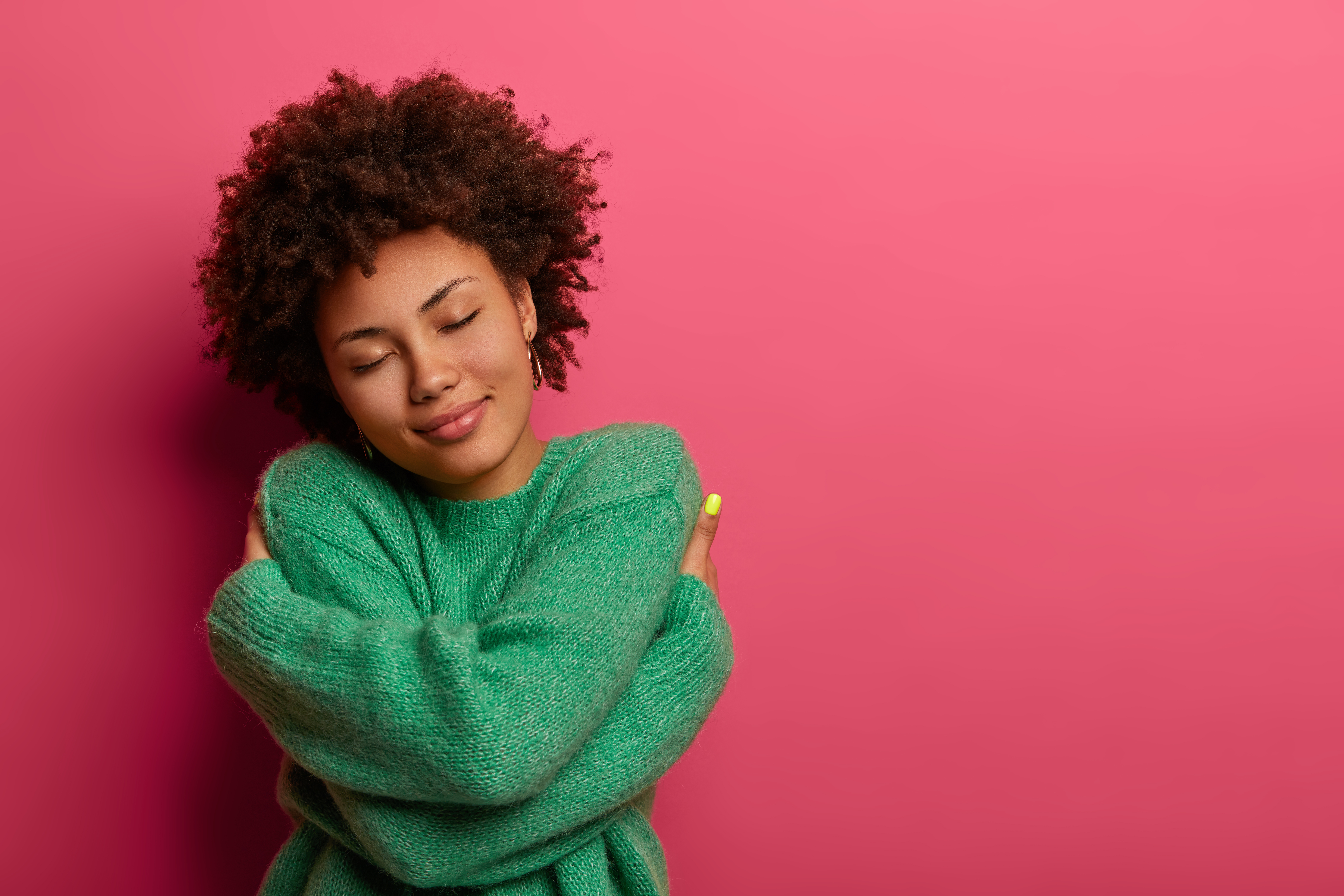 Cheerful young woman expresses self love and care, tilts head and smiles gently, wears green oversized jumper, embraces own body, closes eyes, stands in studio against pink background