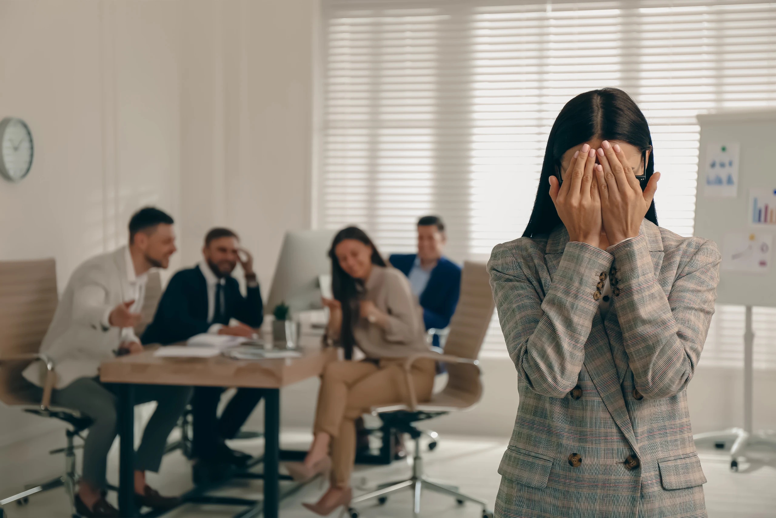 Workplace setting with a distraght and embarassed employee in the foreground while four employees laugh in the background